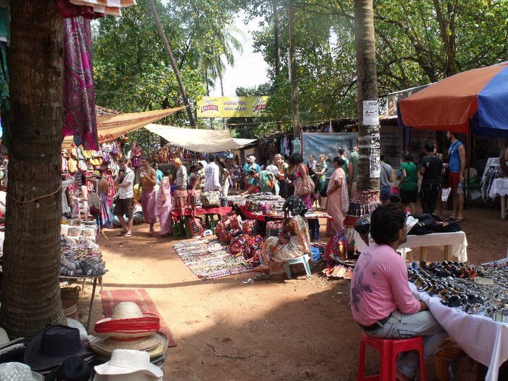 A bustling outdoor market in Goa, filled with vibrant stalls showcasing crafts, clothing, and accessories, surrounded by crowds of people exploring and shopping.
