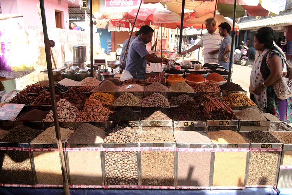 A bustling spice market stall in Mapusa, Goa, showcasing various spices in clear containers while shoppers engage with the vendors.