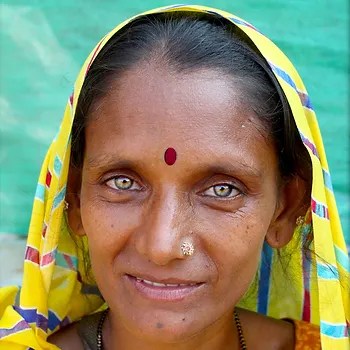 A woman with striking green eyes and a yellow patterned scarf smiles warmly against a colorful background.