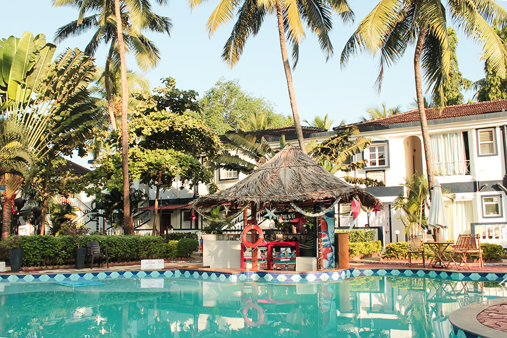 A serene view of Santana Beach Resort in Goa, featuring a swimming pool surrounded by tropical plants and palm trees, with a thatched-roof bar and nearby accommodations.