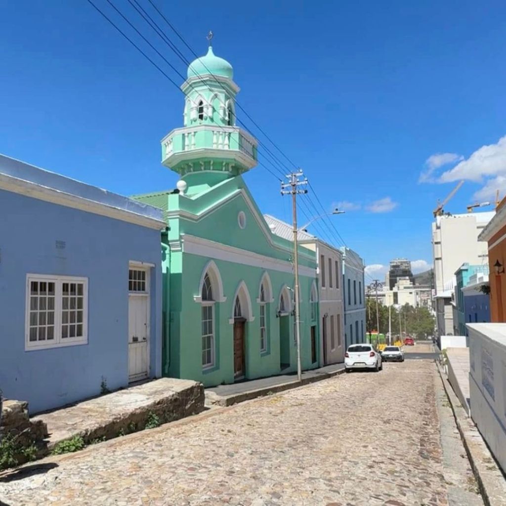 A colorful street in Cape Town featuring a mint green building with a dome and a white facade next to a blue building, under a clear blue sky.