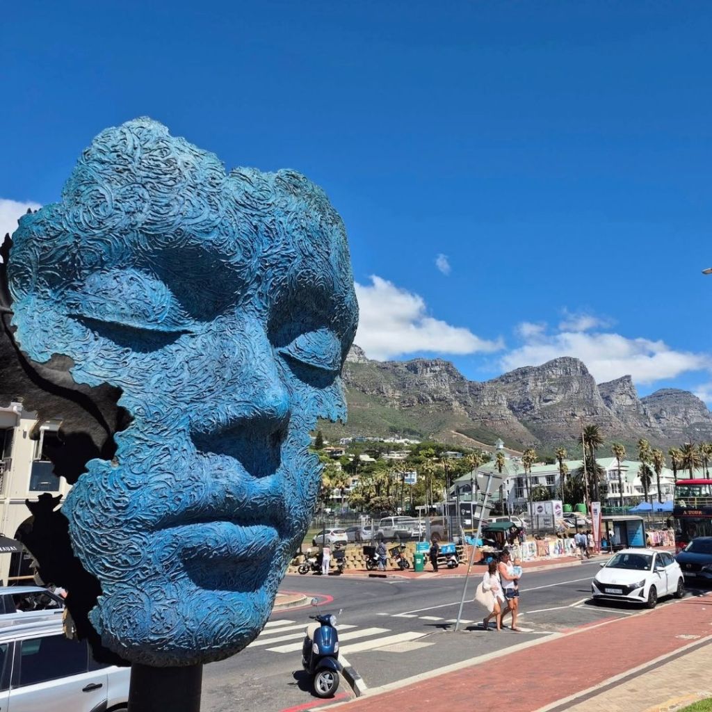 A blue sculpture resembling a face with closed eyes, set against a backdrop of mountains and a clear blue sky in Cape Town.