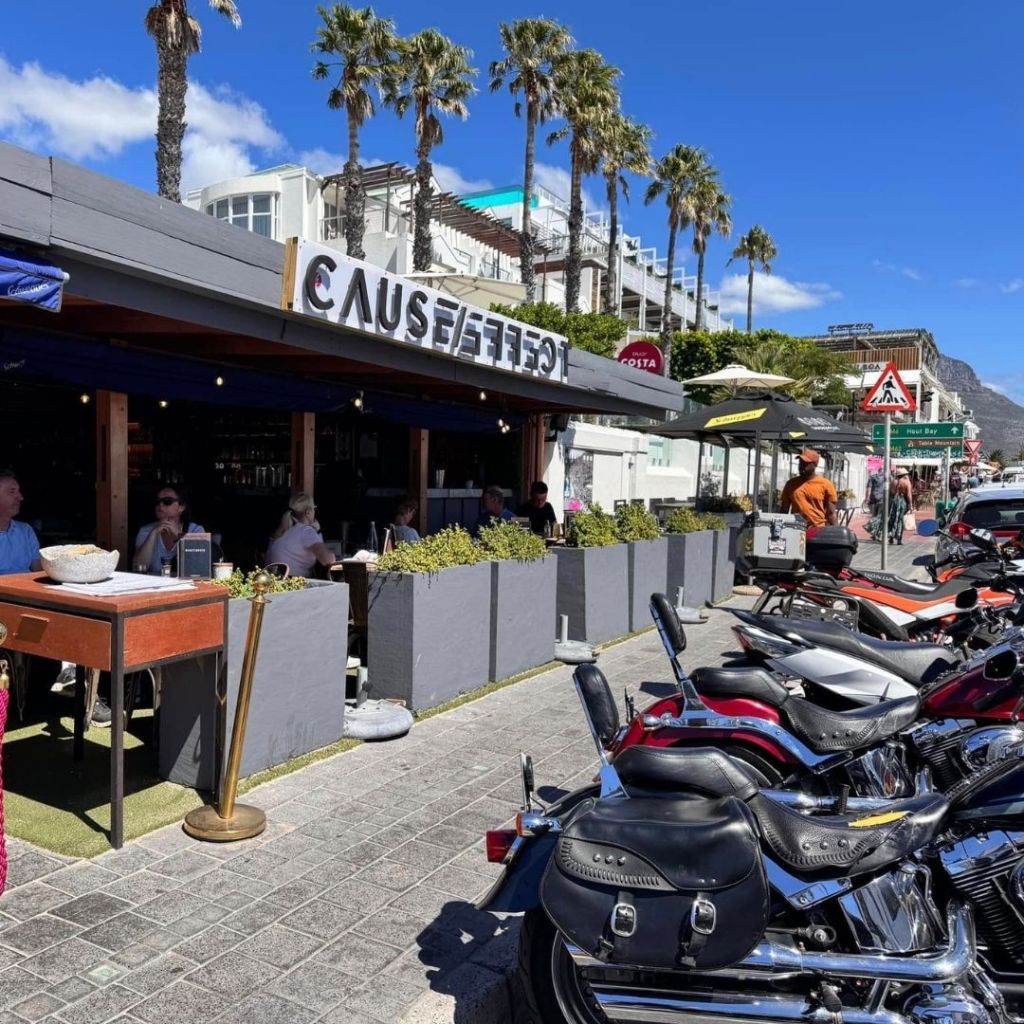 Exterior view of a restaurant named 'Cause Effect' with outdoor seating shaded by umbrellas and palm trees in the background, along a busy street in Cape Town. Several parked motorcycles are visible in the foreground.