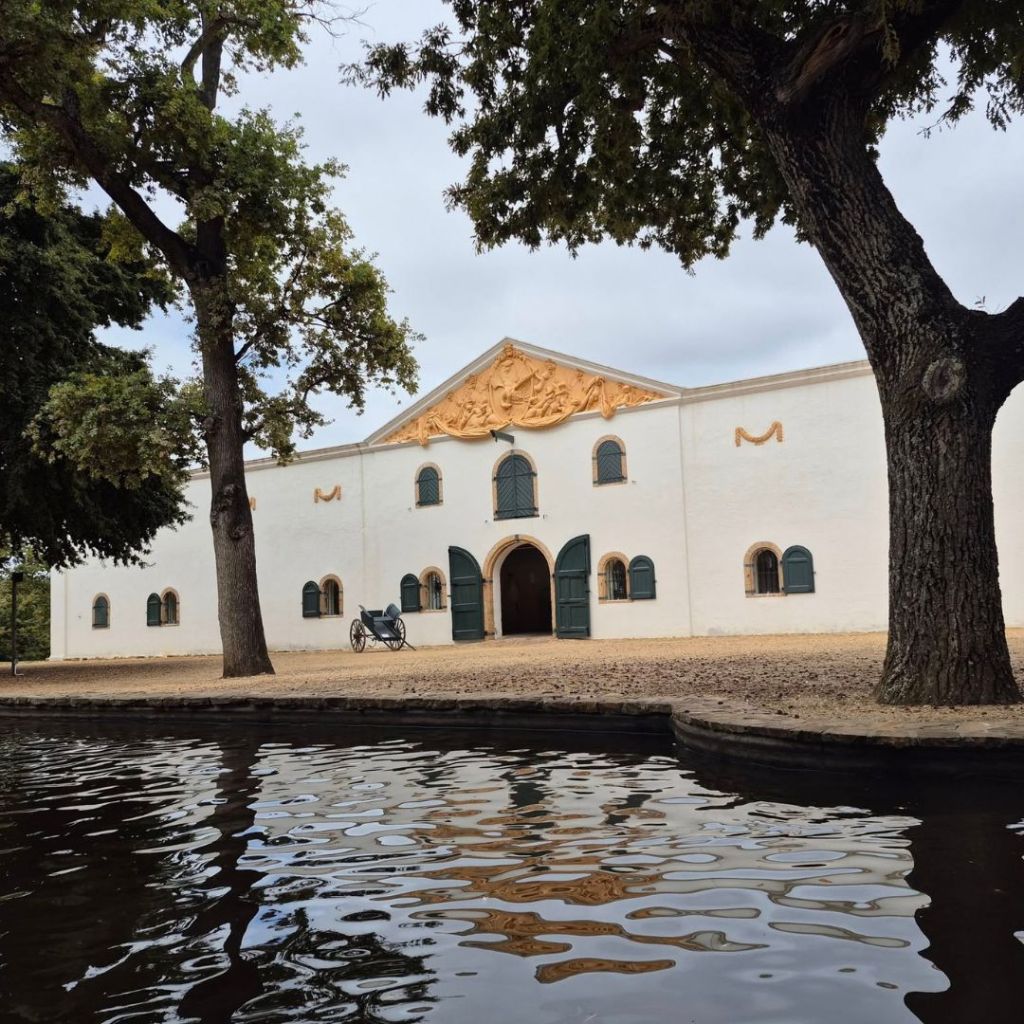 The historic Groot Constantia Manor House, viewed beside a calm body of water, surrounded by trees.