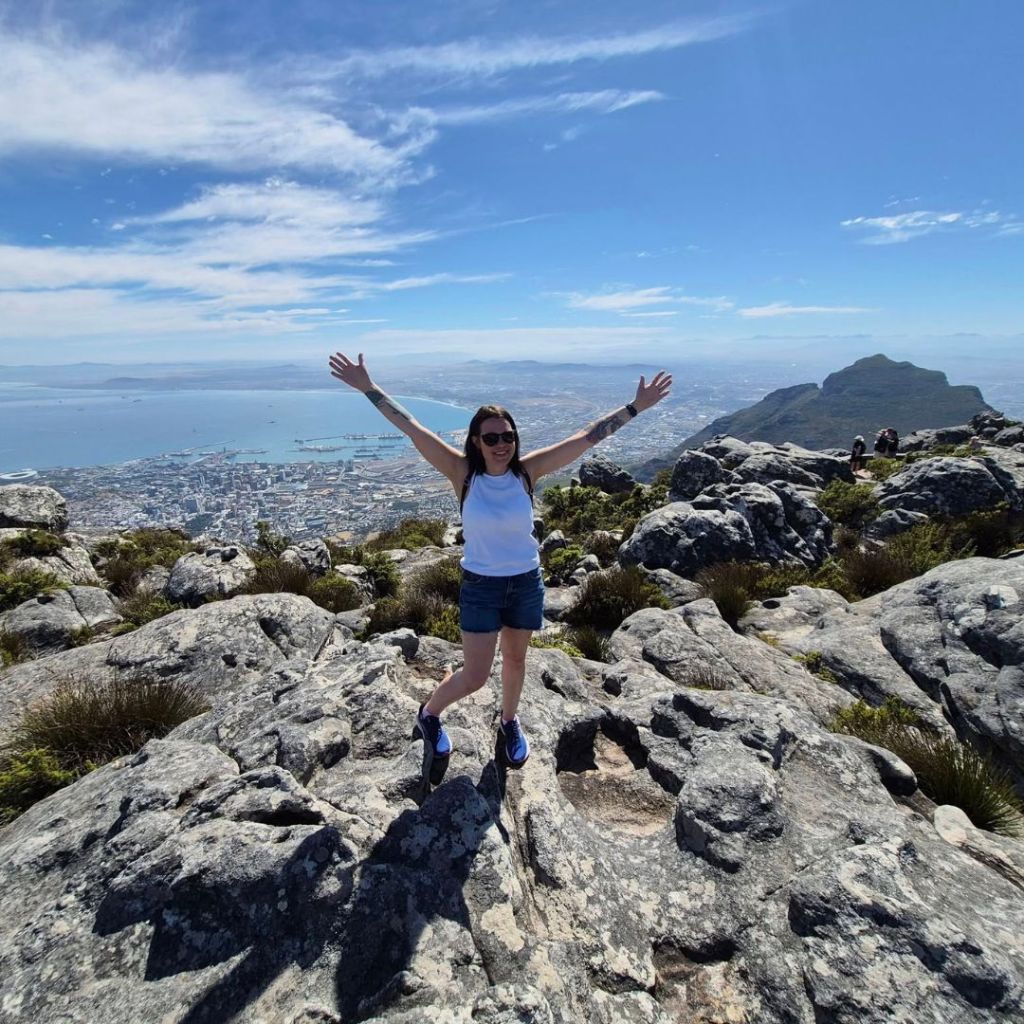 A person standing on rocky terrain at the top of a mountain, with arms raised joyfully, overlooking a scenic view of Cape Town and the ocean below.