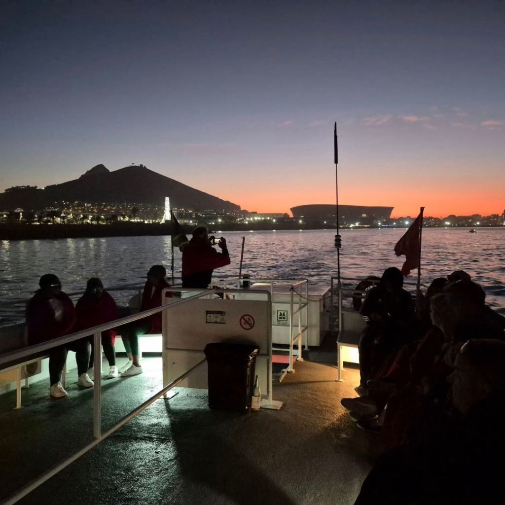 A sunset cruise on a boat in Cape Town, with Table Mountain in the background and vibrant sky colors reflecting on the water. Passengers are seated on the deck, and a crew member stands with a camera.
