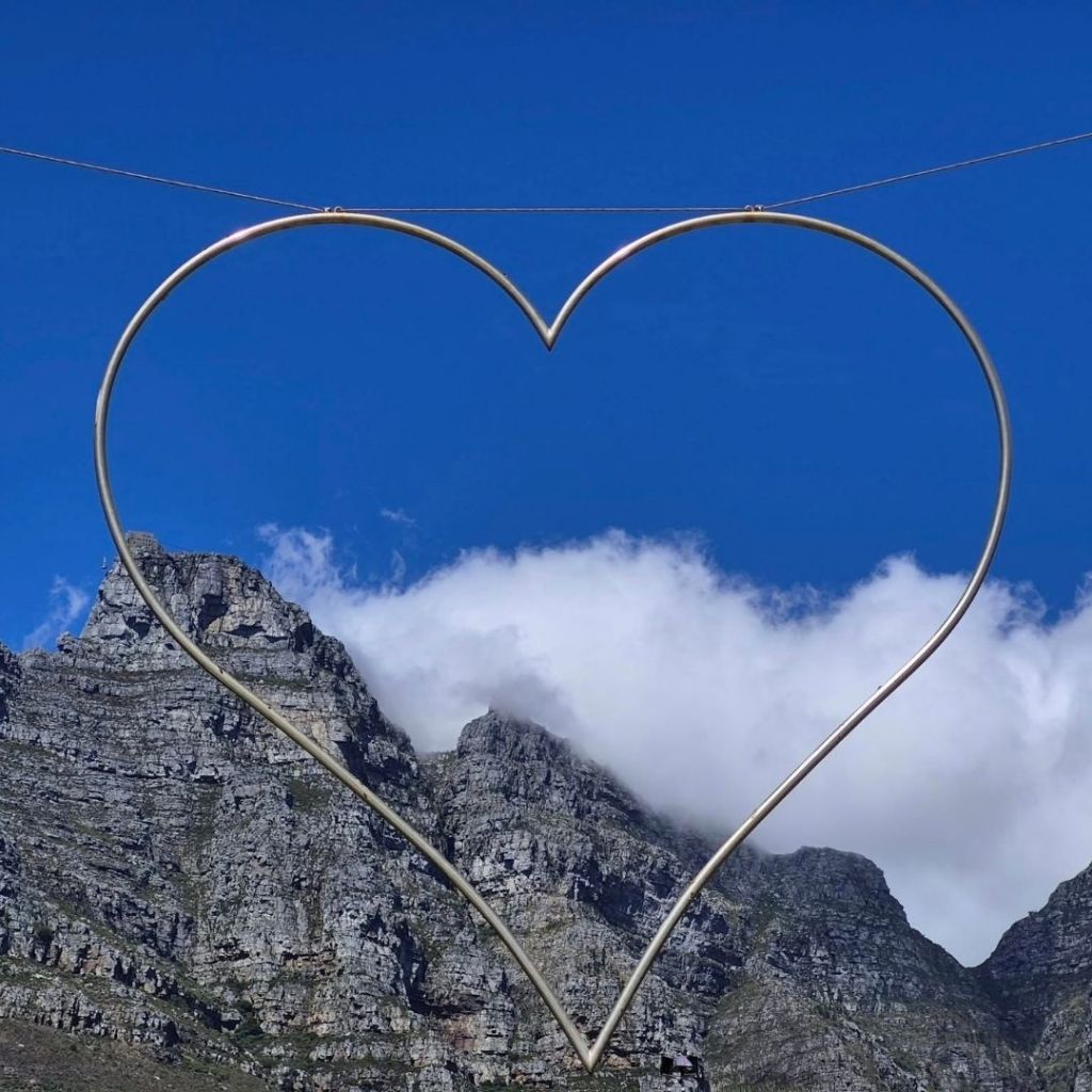 A heart-shaped frame suspended against a backdrop of Table Mountain, with clouds hovering around the mountain peaks under a blue sky.