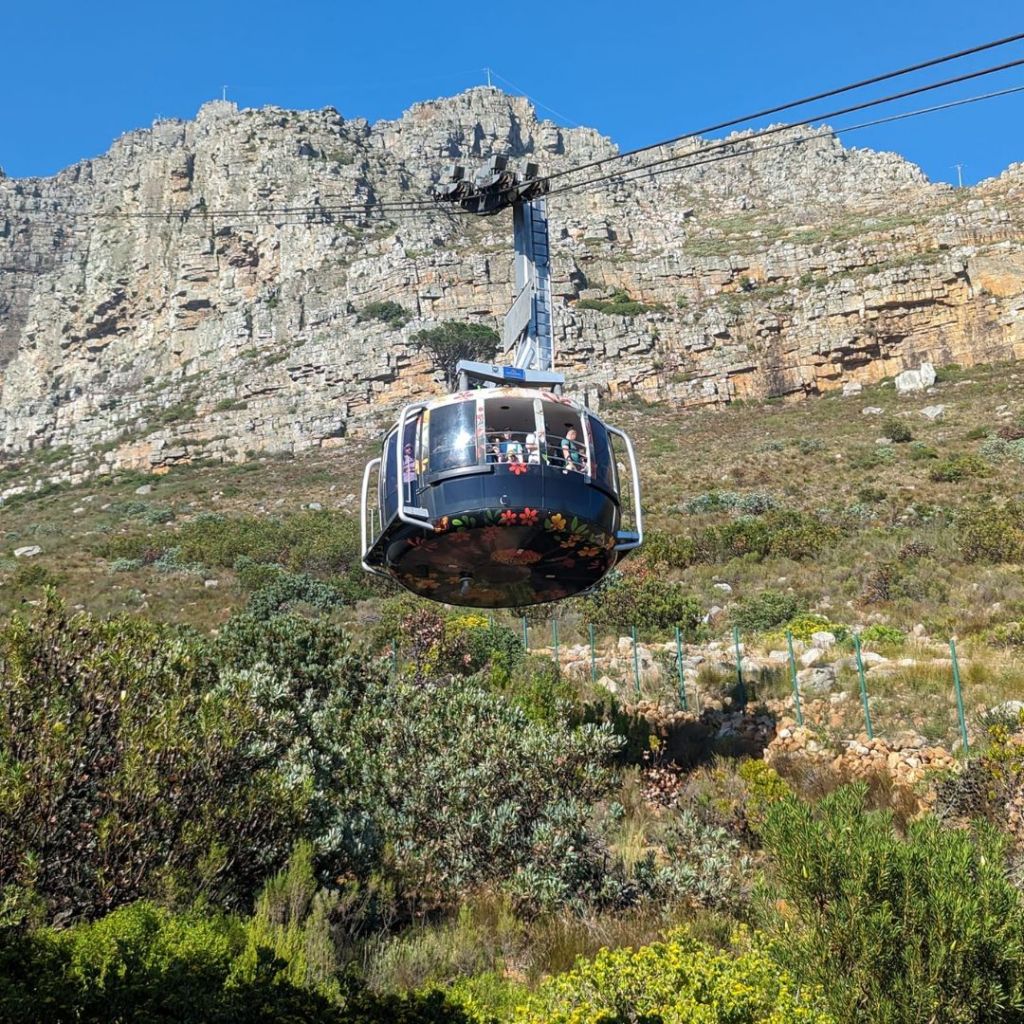 Aerial cable car ascending Table Mountain with passengers inside, surrounded by rocky cliffs and lush greenery.