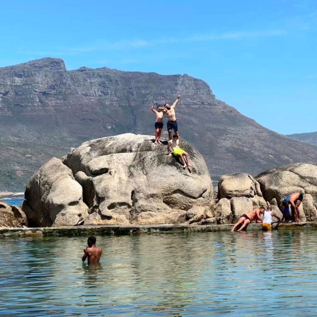 A group of people enjoying their time at a tidal pool, with some climbing on rocks while others swim in the water, set against a backdrop of mountains and clear blue skies.