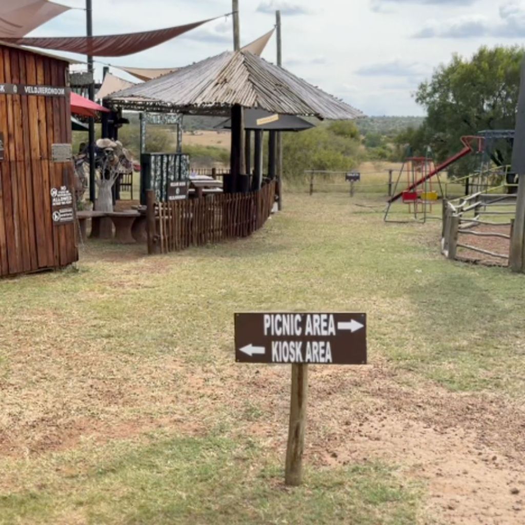 A picnic area signpost directing towards the kiosk and picnic spots at Veldjierondom Picnic Site in Dinokeng Game Reserve, with rustic seating structures in the background.