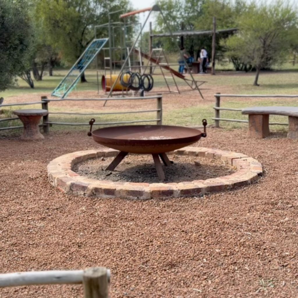 A rustic braai fire pit surrounded by a circular stone border, set in a picnic area with a playground in the background, featuring slides and swings against a natural landscape.