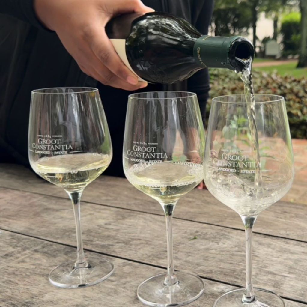 A person pouring wine from a bottle into three glasses with 'GROOT CONSTANTIA' branding on a wooden table.